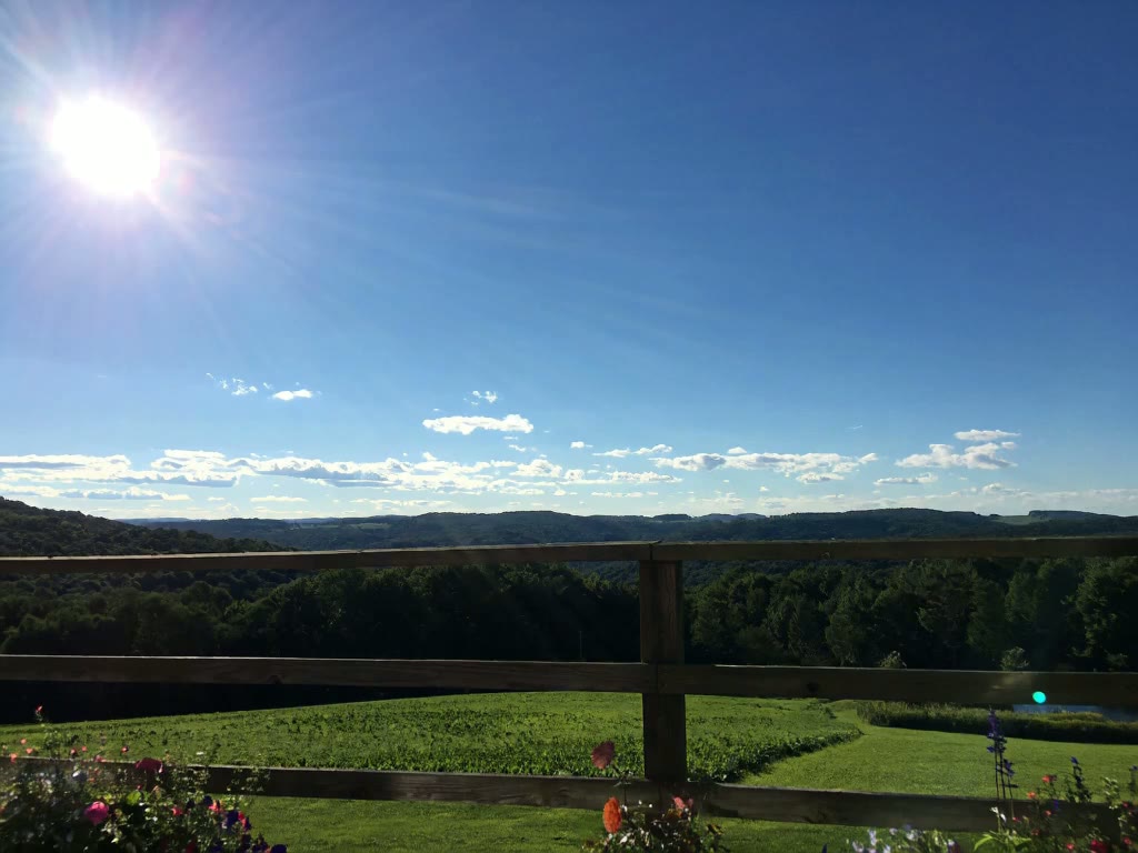 Brilliant sun over a wide Catskills valley, seen past a wooden rail fence with rolling green fields and wooded ridgelines beyond