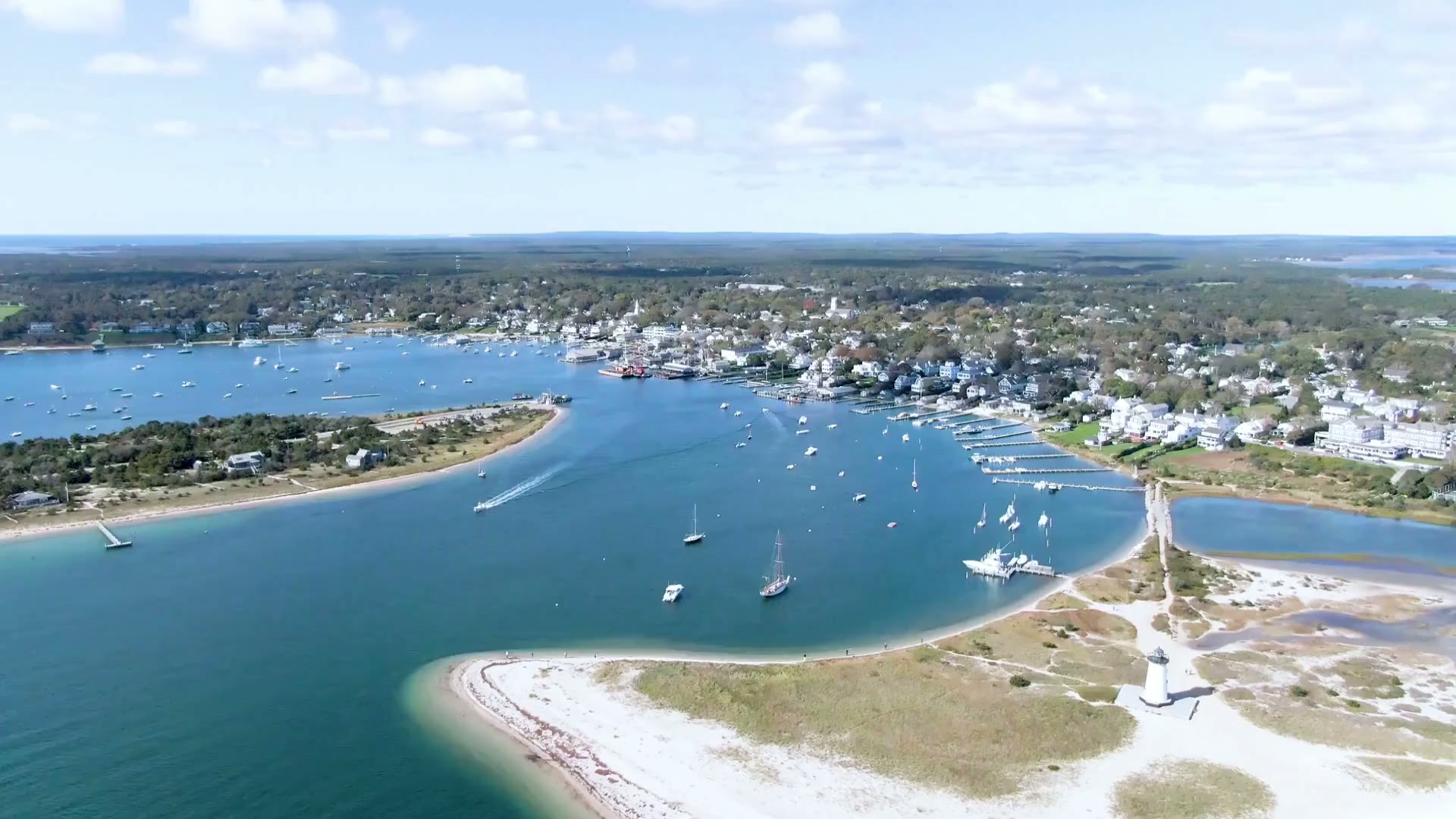 Aerial over a sunlit harbor with sailboats, a white-sand point and a lighthouse